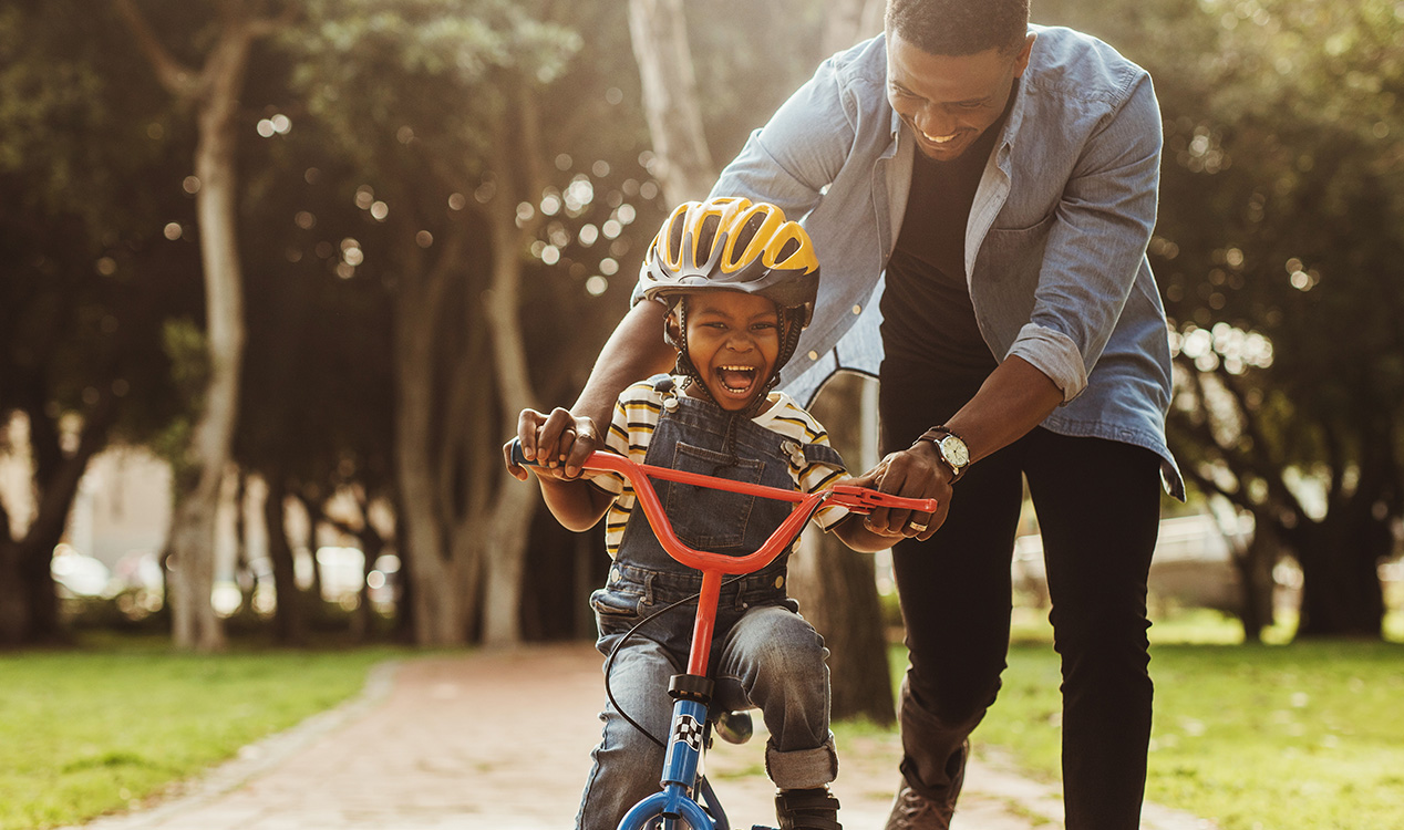 An adult walks beside a young child riding a bicycle, holding the handlebars for support