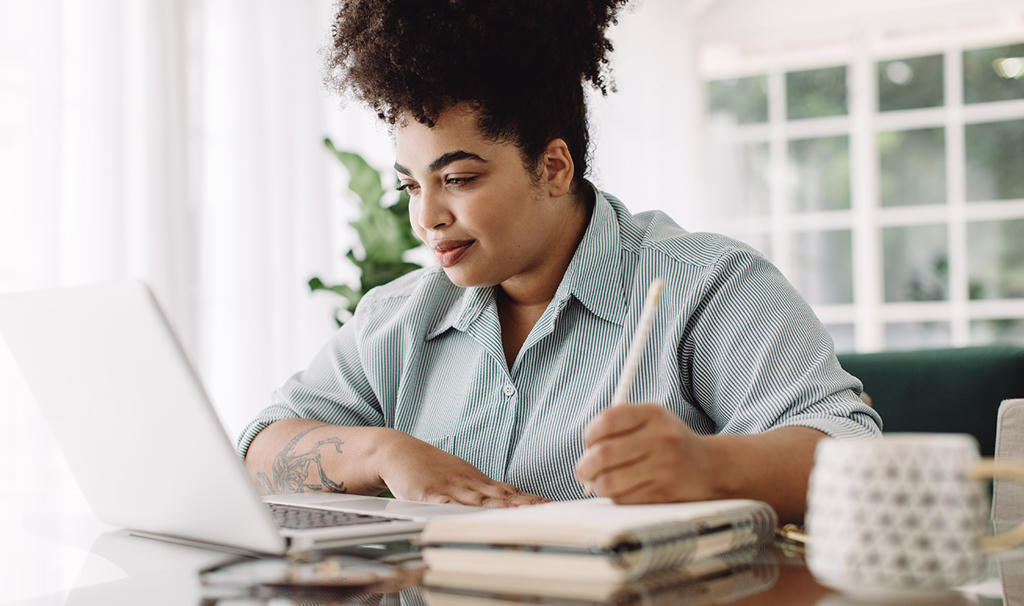 Woman seated at a desk using a laptop while writing in a notebook with a pencil