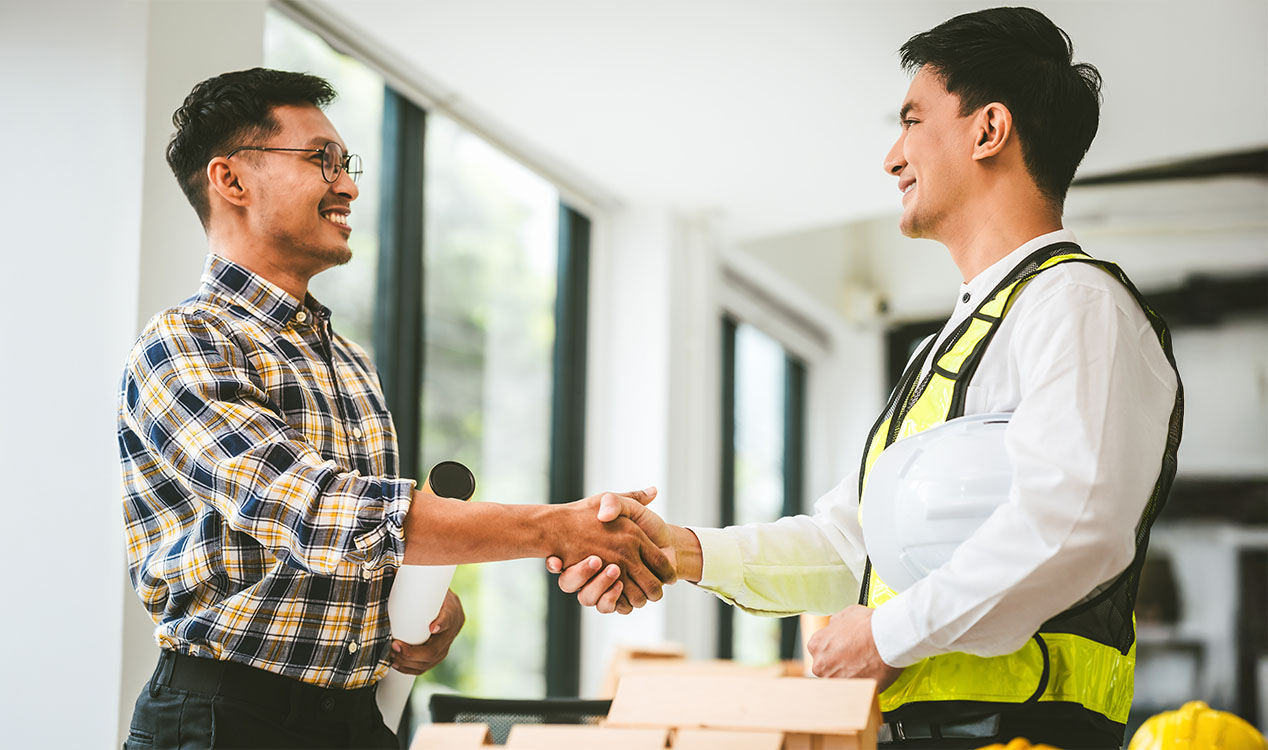 Two professionals shake hands at a construction site, one wearing a safety vest and holding a hard hat.