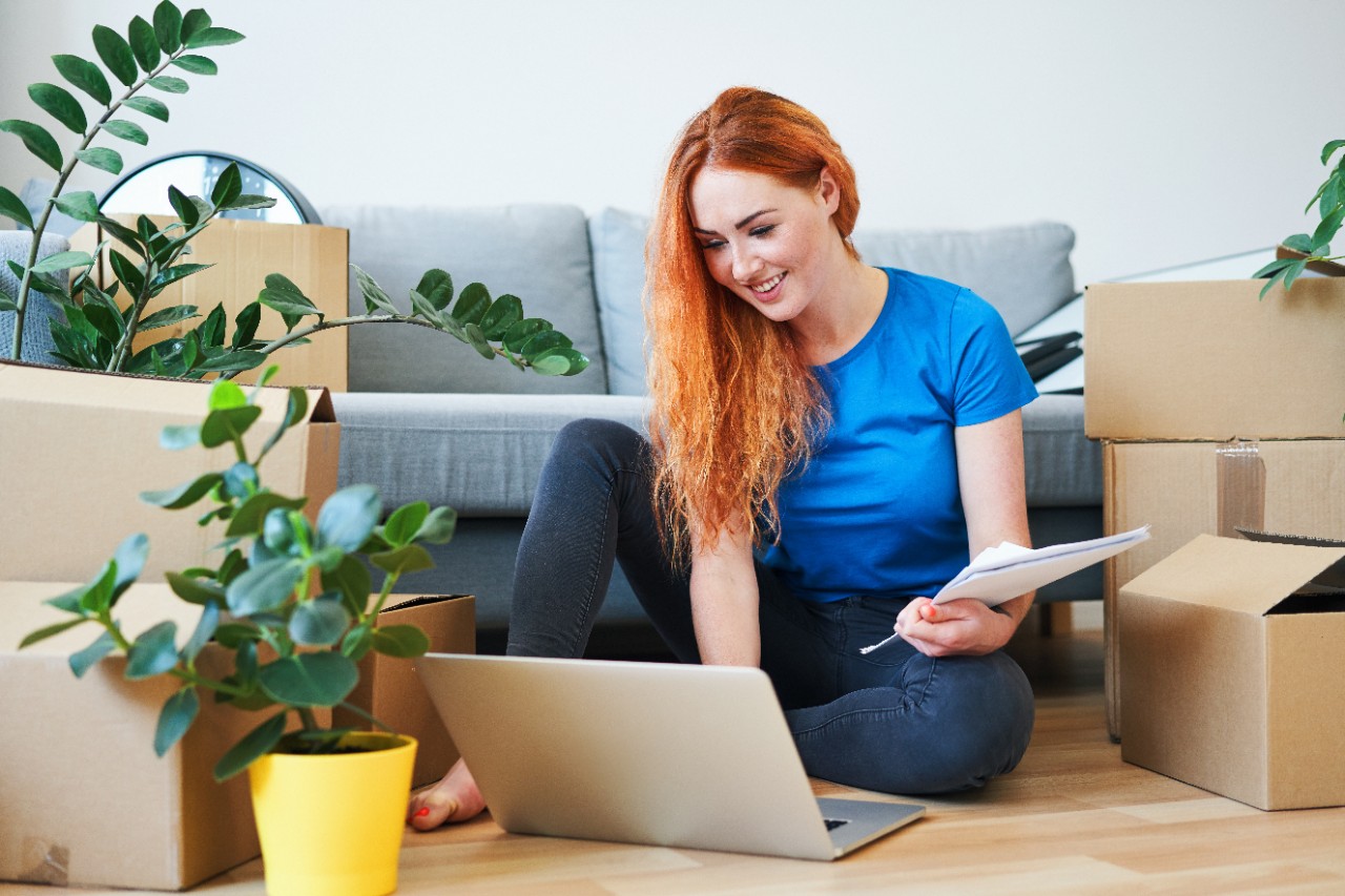 Happy young woman paying bills with laptop while sitting on floor in new apartment