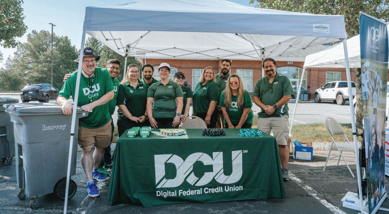 Group of volunteers in matching green “DCU Gives” shirts stand behind a table under a pop-up tent outdoors, with a DCU-branded tablecloth, small giveaway items, a banner, parked cars, and a brick building in the background.
