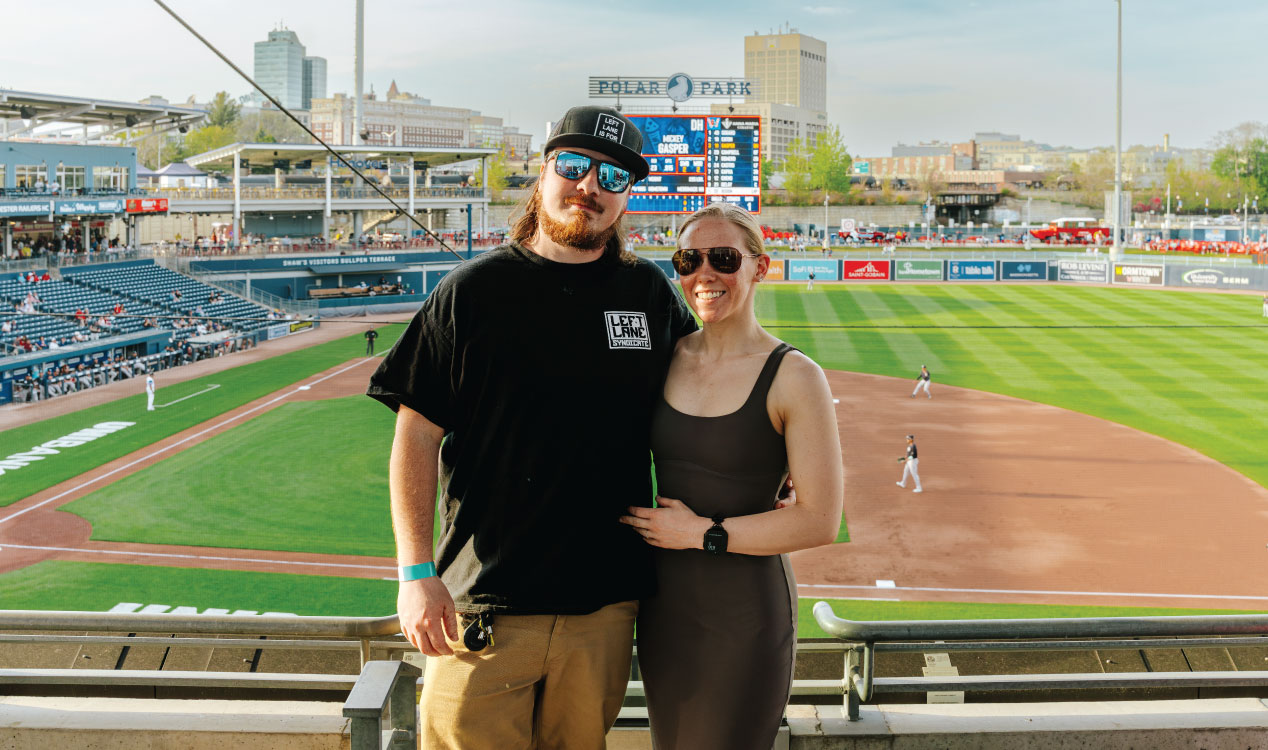 Two people stand together at a stadium railing overlooking a baseball field at Polar Park, with players on the field, seating areas, a scoreboard, and city buildings visible in the background.