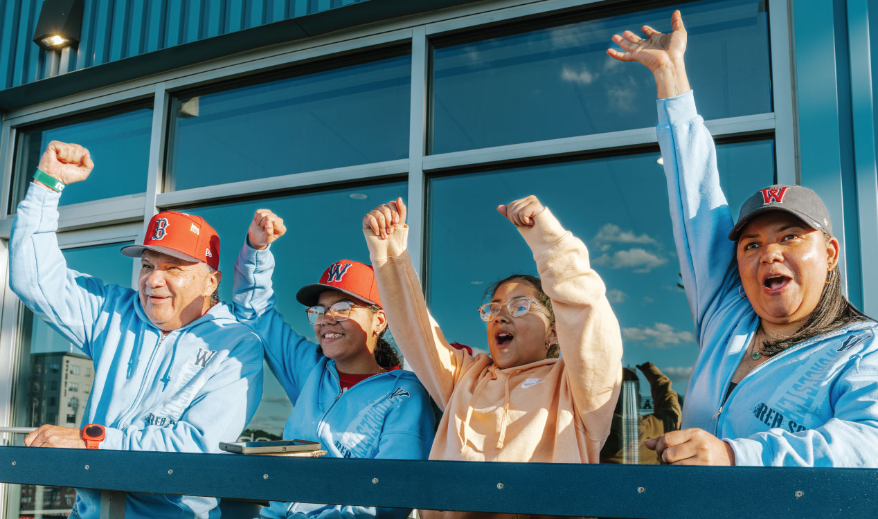 Four people in casual clothing and baseball caps stand at a railing outside a building with large windows, raising their arms in celebration, as sunlight reflects off the glass behind them.