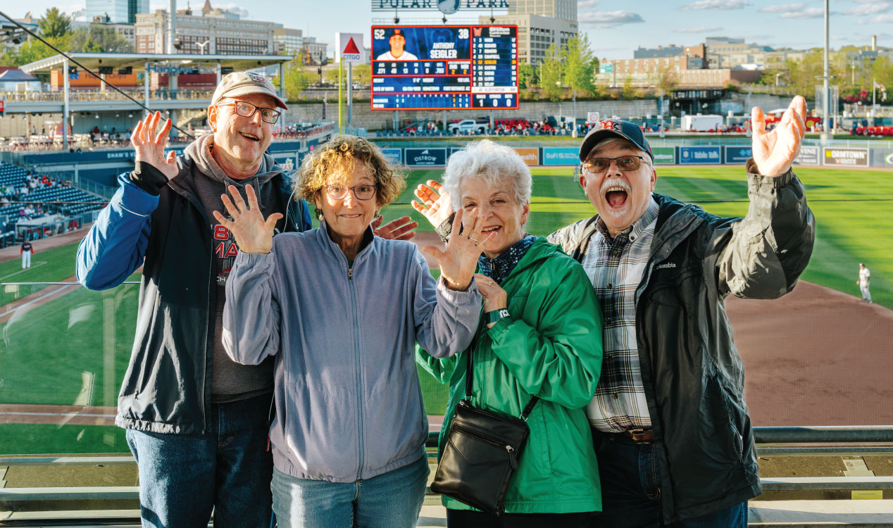 Four adults stand at a railing at Polar Park, waving toward the camera with a baseball field, scoreboard, and stadium seating visible behind them on a sunny day.