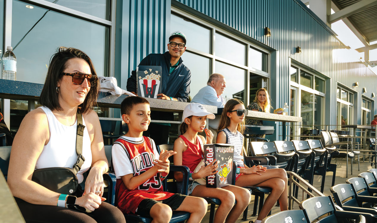 Four people and three children sit in stadium seats at a baseball game, with popcorn and drinks, while others stand behind a railing; a modern building with large windows and rows of empty seats extend along the right side.