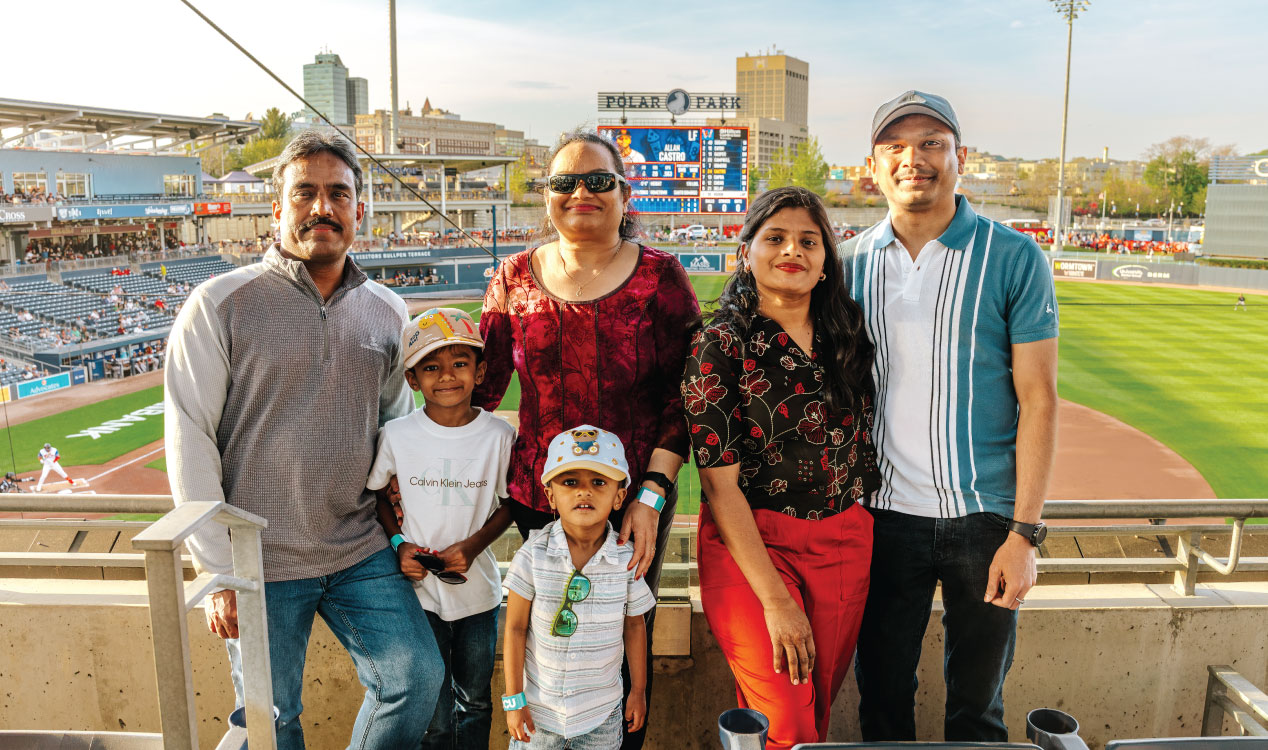 Six people, including two children, stand at stadium seating railing at Polar Park with a baseball field, scoreboard, and city buildings in the background on a sunny day.