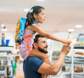 Person carrying a child on their shoulders while holding an object in a store aisle, with school supplies visible on nearby shelves.