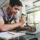 Student holding a coffee while working on a laptop with a notebook by the side