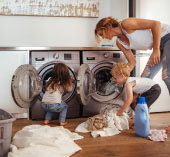 Two people loading towels into front-loading washing machines in a laundry room, with detergent bottles and folded linens on the floor nearby.