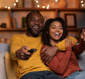 Two people sitting on a couch in a living room, holding a TV remote and taking a selfie together, with warm lighting and shelves in the background.