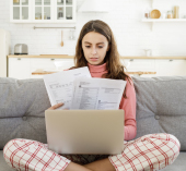Person sitting cross-legged on a couch with a laptop and reviewing printed bills or financial documents in a bright home setting.