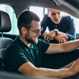 Man sitting behind the steering wheel of a car while another man leans in from the open drivers-side window