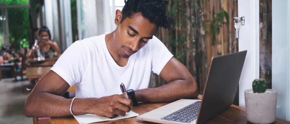 Man sitting at a table and taking notes on paper from his laptop.