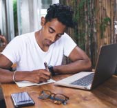 Man sitting at desk with calculator and laptop, writing out financial goals