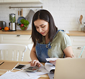Woman standing in kitchen with laptop, calculating interest on saving accounts vs. checking