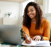 A woman on her laptop while writing in a book.