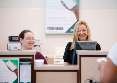 Two DCU branch employees behind the teller desks smiling. 