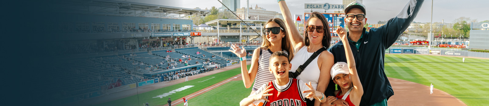 Group of five people in stadium seats at Polar Park pose and raise their arms, with a baseball field, scoreboard, and city buildings visible in the background.
