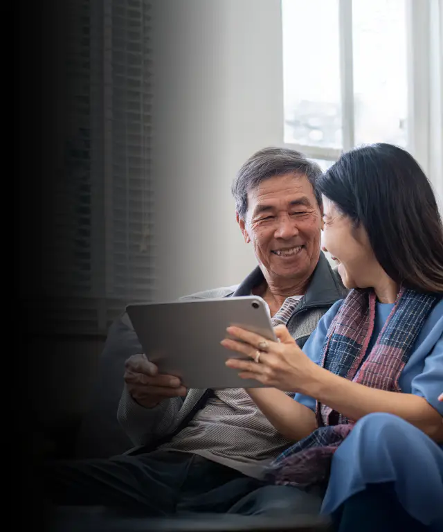 Two people sitting together on a couch in a bright living room, holding a tablet and looking at the screen, with plants and window blinds in the background.