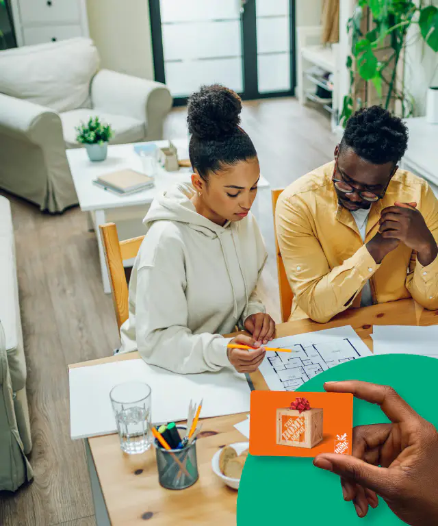 Hand holding a Home Depot gift card in front of a couple reviewing their home floor plan together while sitting at a table.