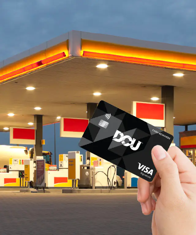 Hand holding a DCU Visa Signature card in front of a brightly lit gas station at dusk, with fuel pumps visible and promotional text reading “Earn 2X Points” in the upper corner