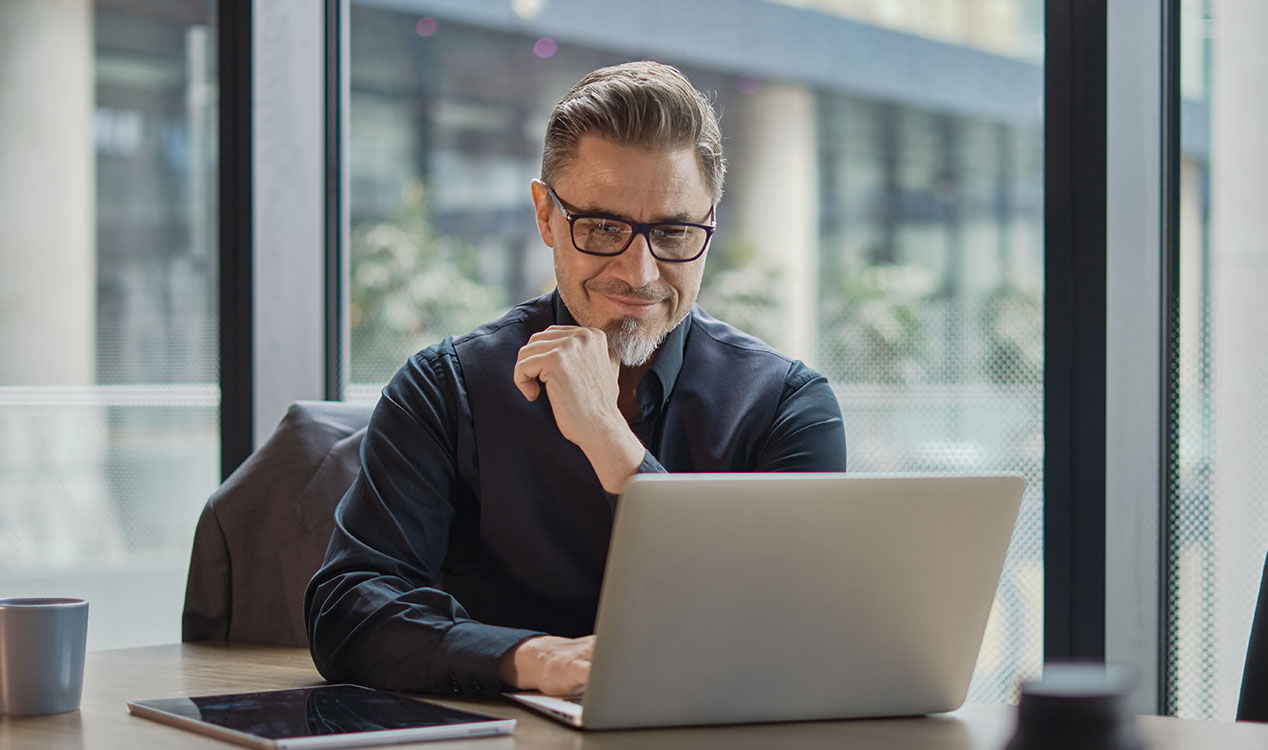 Business professional wearing glasses sits at a desk using a laptop, with a tablet and coffee nearby in a bright office