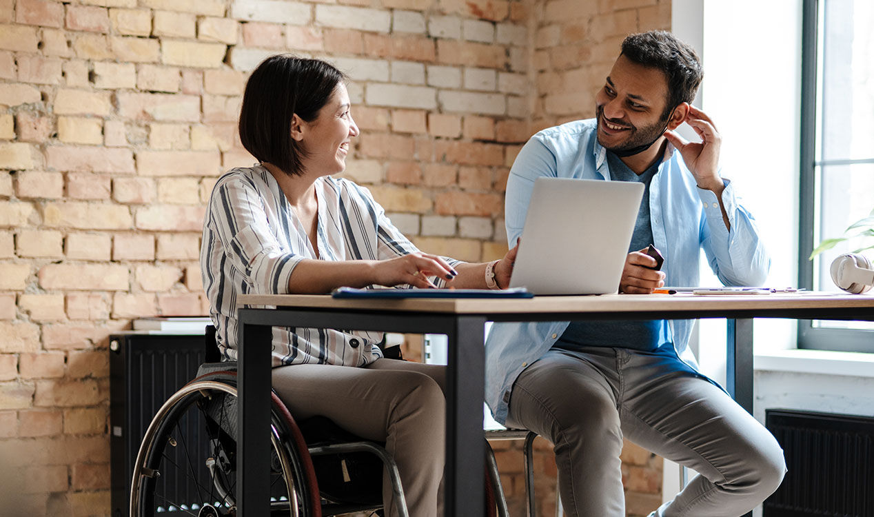 a woman in a wheelchair using a laptop at the desk with a man sitting next to her