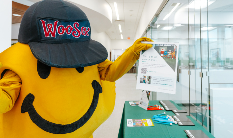 WooSox mascot Smiley Ball holds a promotional poster about the WooSox DCU Member Appreciation Sweepstakes inside one of DCU's three Worcester branches.