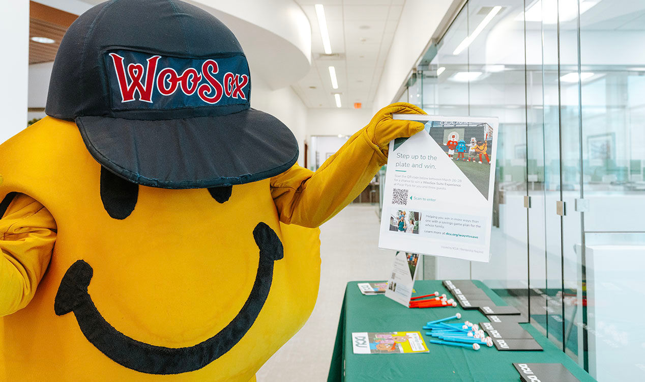 WooSox mascot Smiley Ball holds a promotional poster about the WooSox DCU Member Appreciation Sweepstakes inside one of DCU's three Worcester branches.