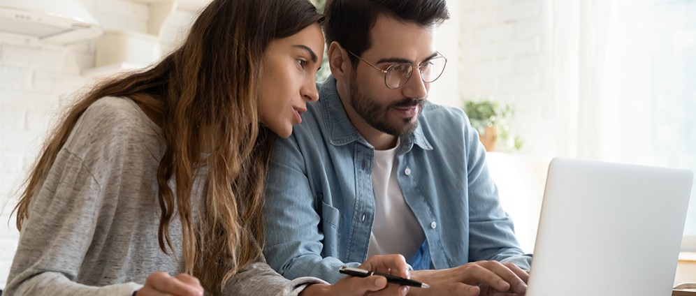 Two people sitting at a table, working together on a laptop while reviewing notes and holding pens.