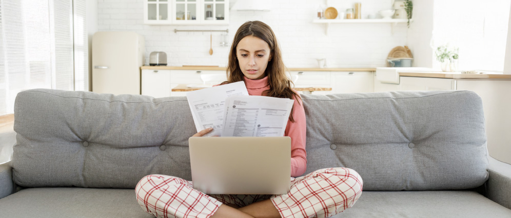 Person budgeting for unexpected expenses at home, sitting on couch with laptop and financial documents in a modern kitchen-living room