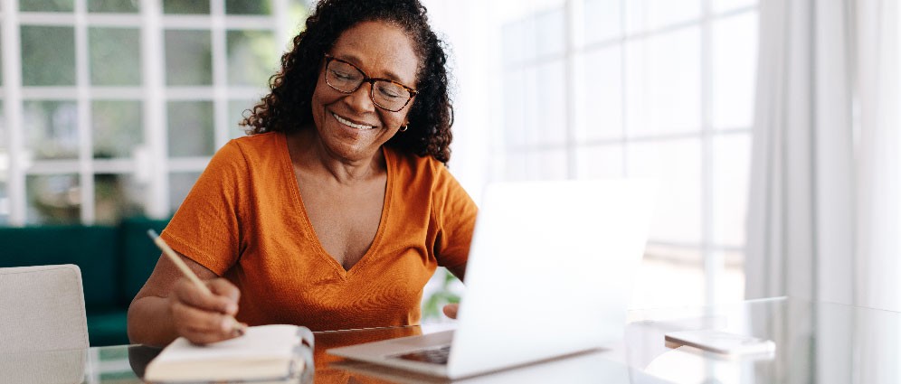 Older woman sitting at her laptop, writing in notebook