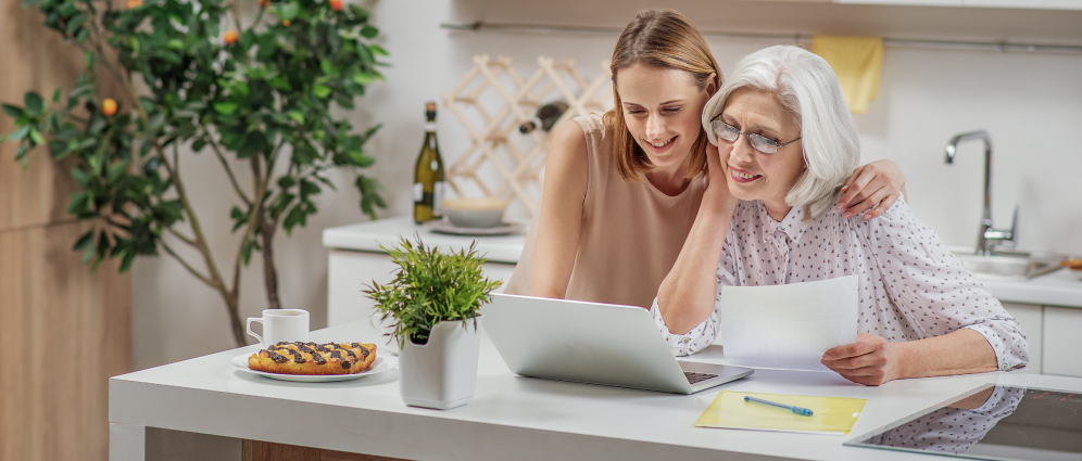 mother and daughter looking at a laptop on a table while the mother holds a paper in her hand
