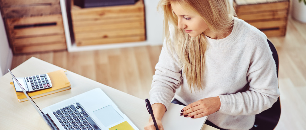 A woman writing at her desk while looking at a laptop